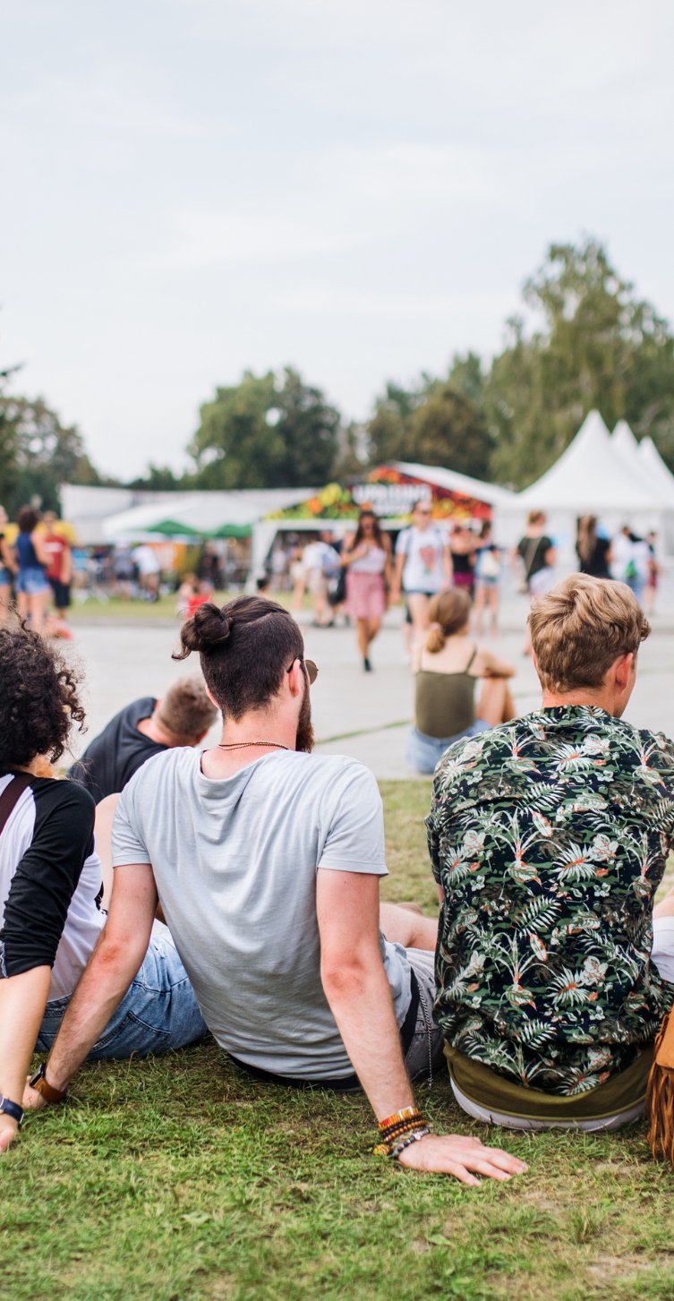 Festivalbesucher sitzen auf Wiese mit Blick auf Essensstände und Zelte Entspannte Festivalatmosphäre – idealer Ort für praktische Give Aways und gebrandete Streuartikel von Firmen.